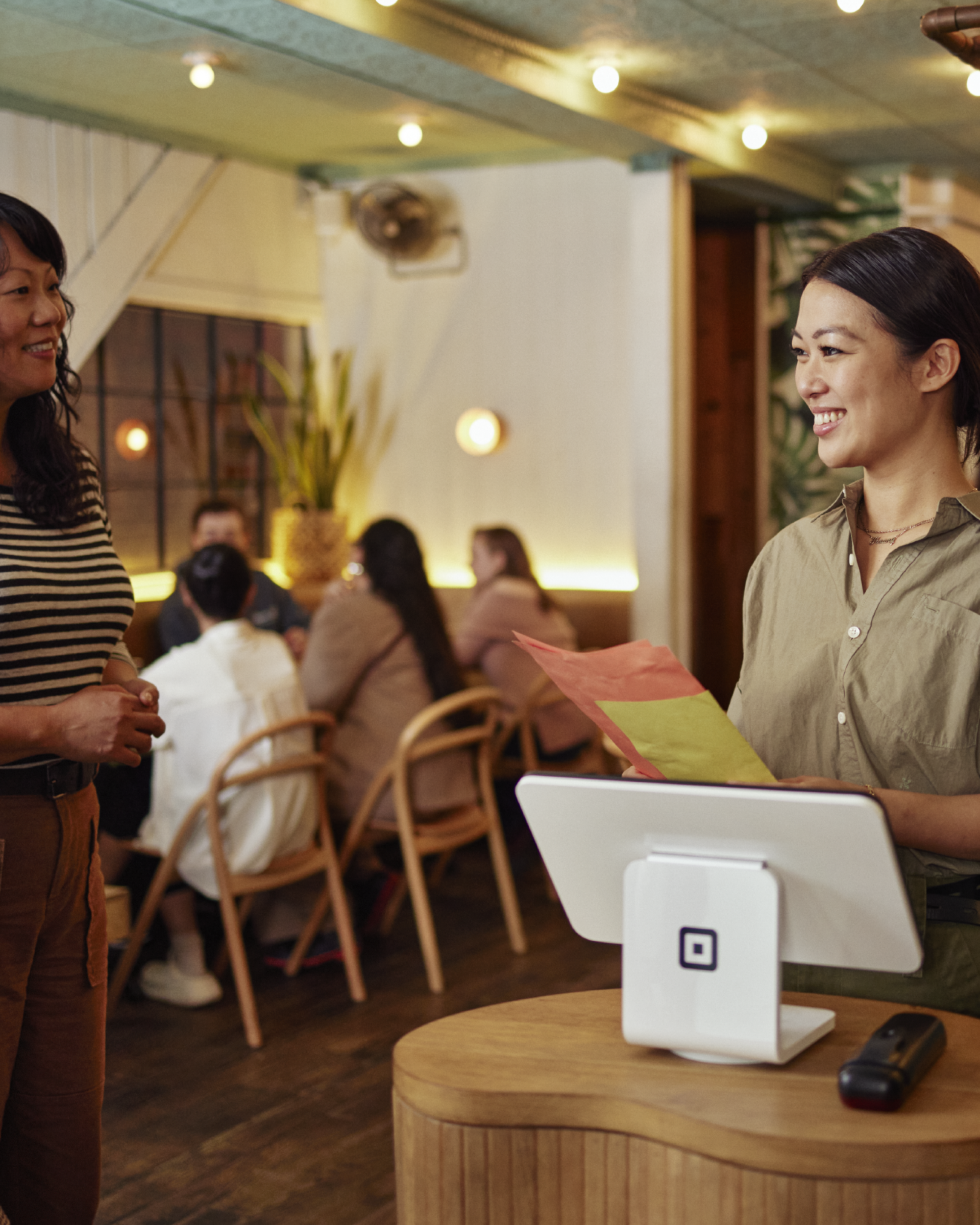 Female hostess inside clean bright modern full service restaurant standing behind Square POS at front desk holding dinner menus and welcoming guest.